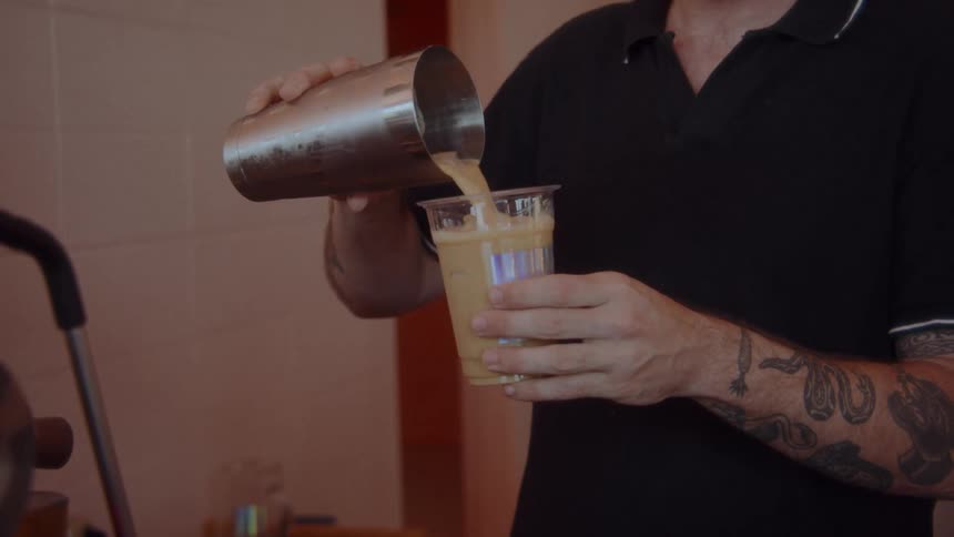 Close up of a young man pouring a drink into a transparent cup for a customer while working at a cafe during daytime