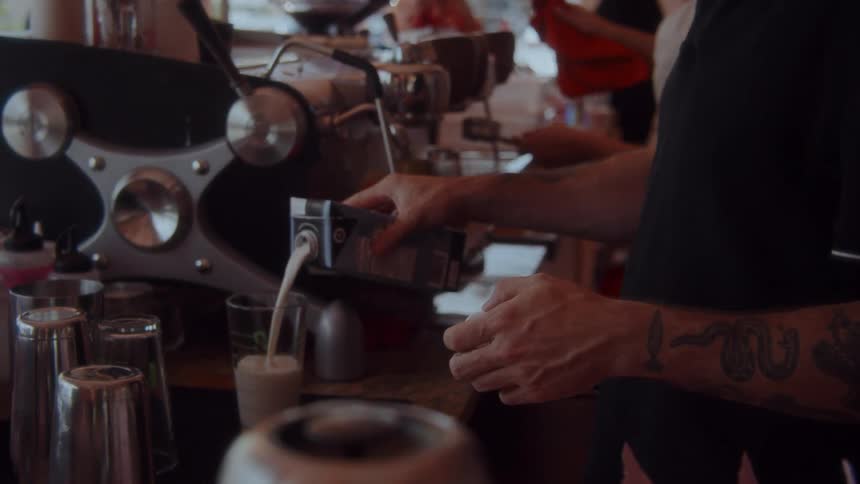 A barista with tattoos pours milk from a carton into a glass at a busy coffee station