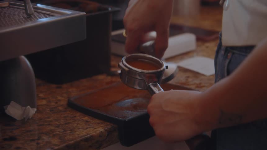 A coffee shop barista uses a tamper to press down on coffee grounds inside a portafilter then locks the portafilter into a large espresso machine with a pressure gauge