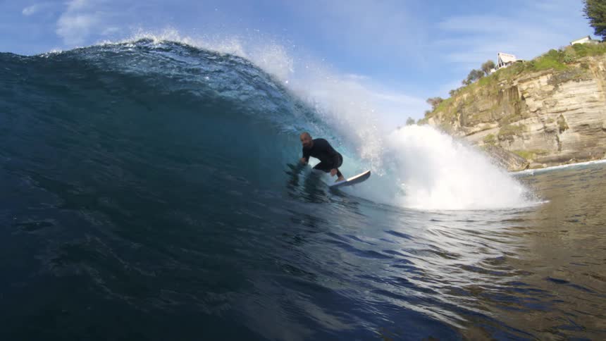 A man stands on a surfboard and rides a wave near a landmass with a cliff and trees