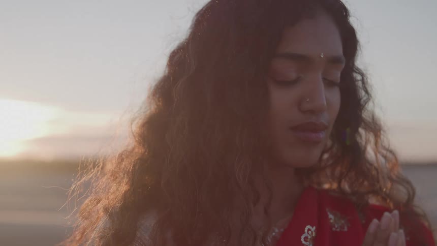 A young woman with long curly hair wearing a red sari stands on a beach with her eyes closed and hands together in a prayerful or meditative pose as the sun sets behind her creating a warm golden flare
