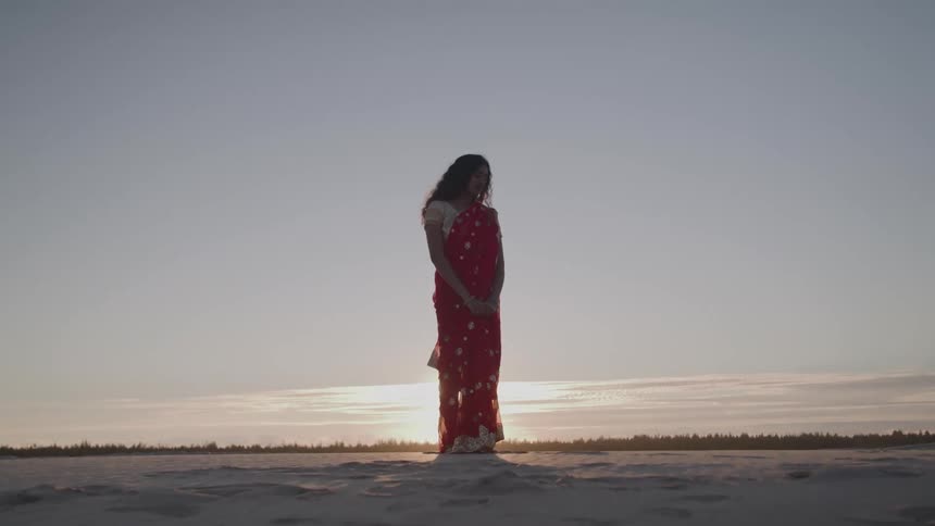 The video shows a woman standing in a large open field with the sun setting behind her silhouetting her figure she is wearing a long red traditionalstyle dress