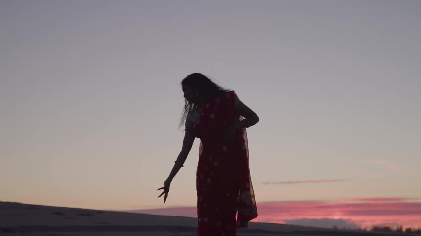 A hindu woman is silhouetted against a sunset sky as she performs a graceful dance on a sandy desert landscape with her arms and hands moving in an expressive way