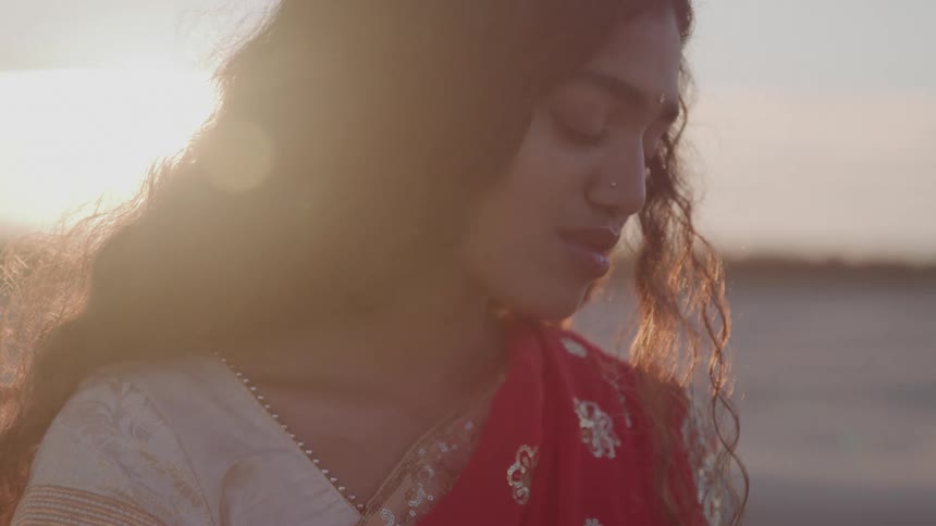A hindi woman with long brown hair and a nose piercing is wearing a red and white patterned garment and looking down at the ground with a soft expression on her face as the bright sun shines behind her