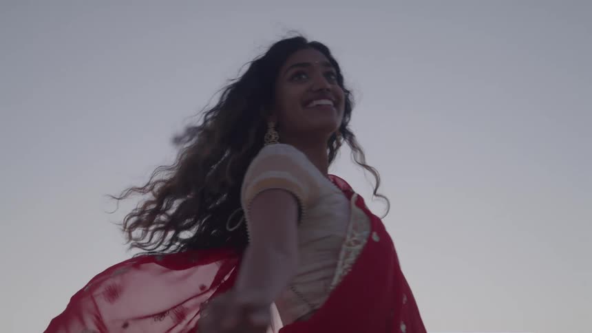 A shot focuses on a person wearing a red and gold sari as she twirls in a field under a clear blue sky the woman is smiling with her hair flowing as she dances