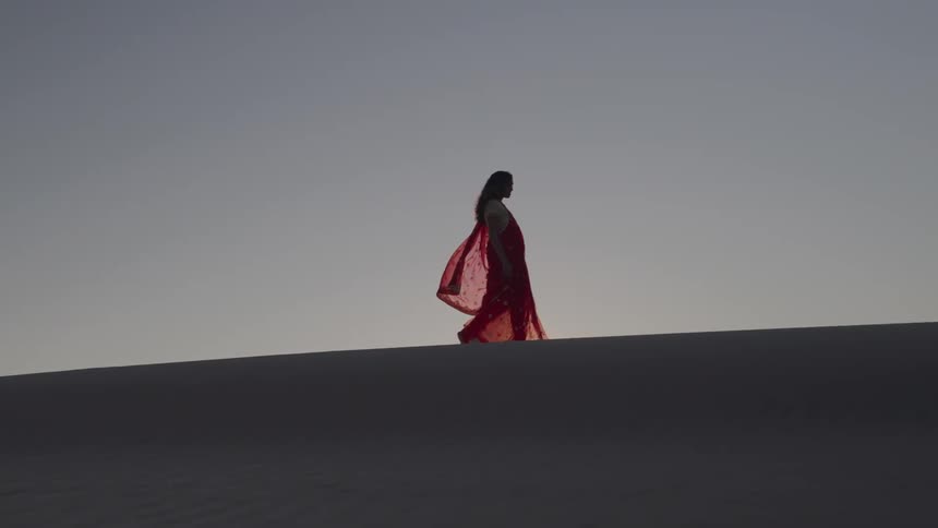 A wide shot shows a woman walking on a ridge in a desert landscape she is wearing a flowing red dress that stands out against the pale sandy terrain