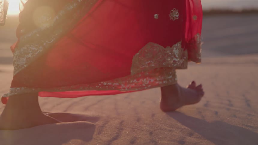 Bare feet walk on the sand with a red dress embroidered in gold