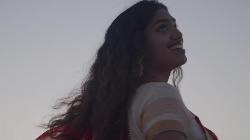 Woman wearing a bodice and a sari spins in place on the beach under a red tinted sky
