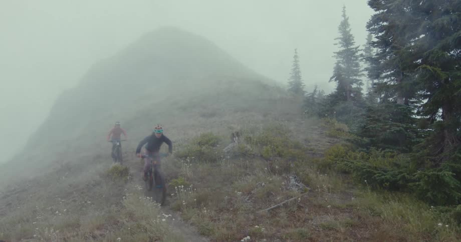 Two people ride mountain bikes along a hilltop trail on a misty day