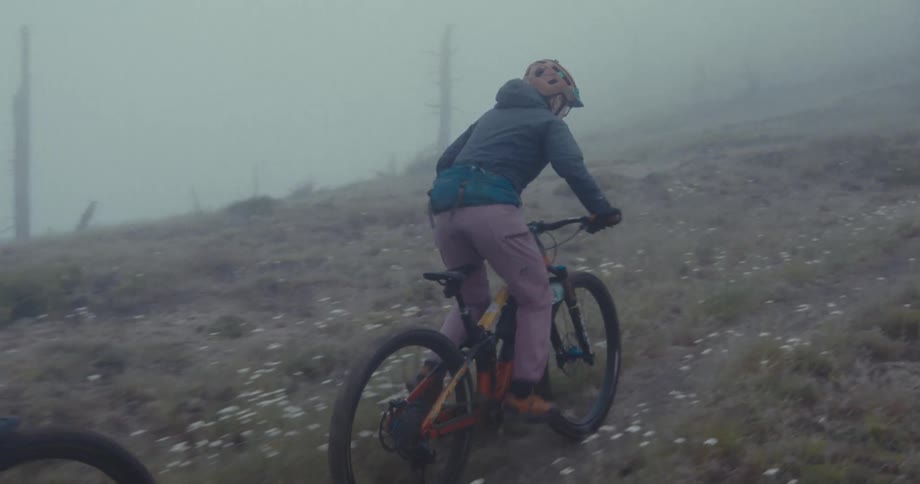 A group of two friends wearing helmets and jackets are mountain biking on a dirt path in a foggy open field with sparse plants