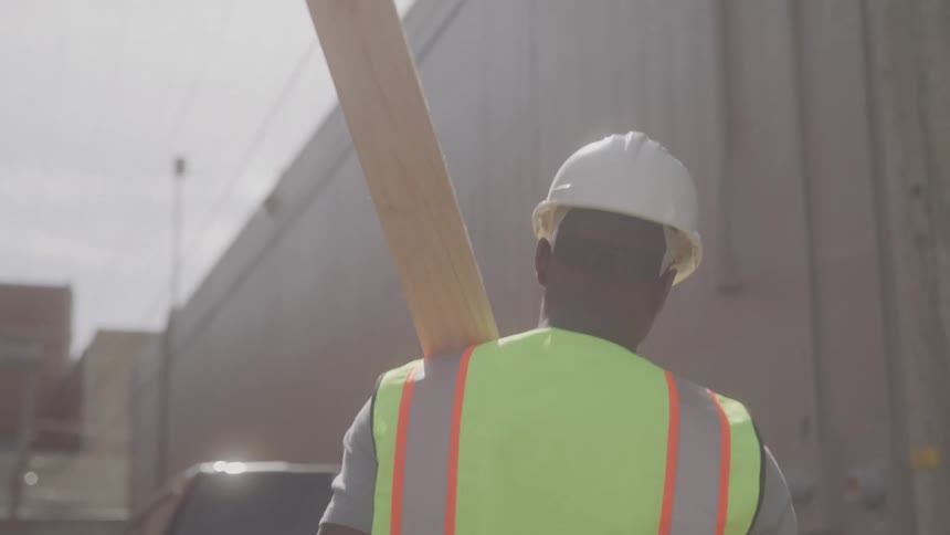 The video shows a construction worker a black man wearing a white hard hat and a yellow safety vest walking away from the camera he is carrying a long wooden beam on his shoulder another construction worker is visible in the background also wearing a