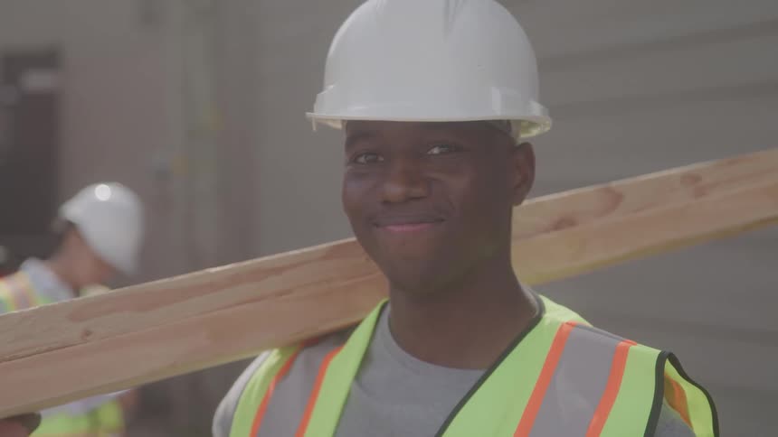 A male construction worker wearing a white hard hat and a fluorescent safety vest carries a wooden plank on his shoulder looks directly at the camera and smiles