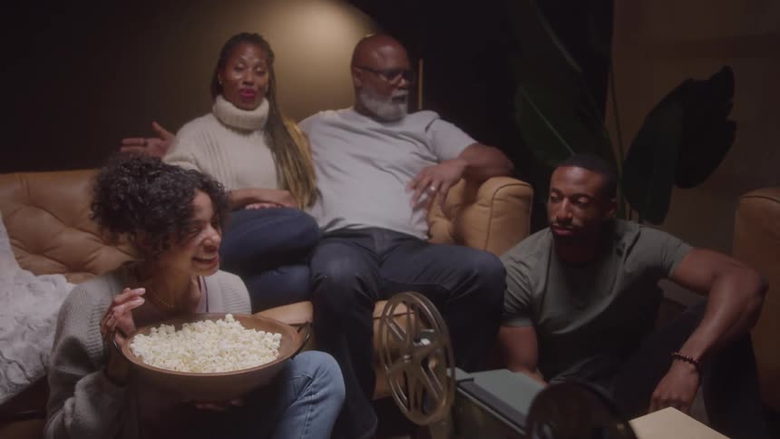 A multigenerational family is gathered in a dimly lit room enjoying a movie night with popcorn they are watching a film projected from a vintagelooking reeltoreel projector