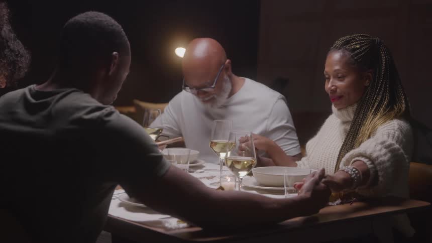 Family holds hands to pray together at a dinner table before the meal