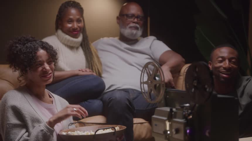A happy family of four including a man a woman a young woman and a young man is gathered around an antique film projector with a large reel and watching a movie while eating popcorn