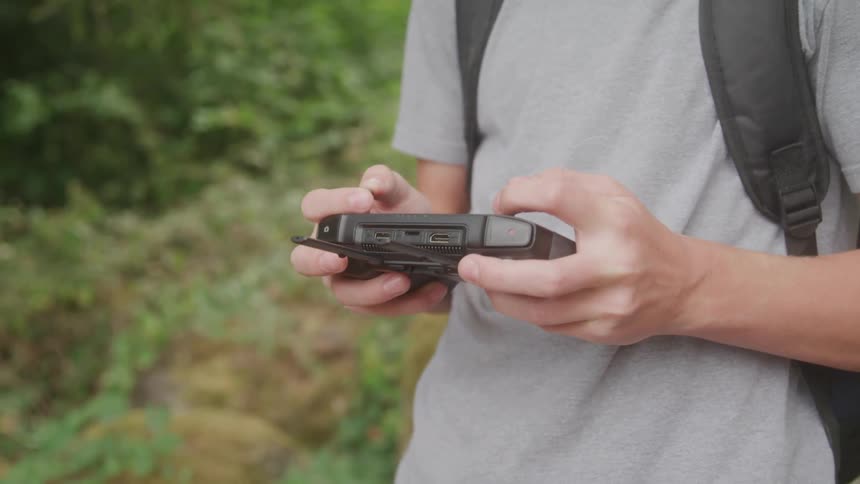 A close view of a persons hands shows them holding a small black electronic device with various ports and buttons while their thumbs press the controls