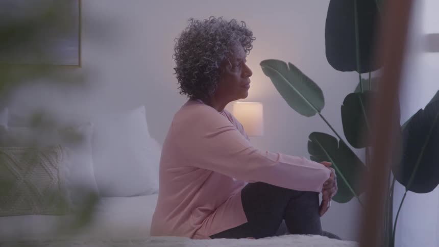 A woman with curly hair sits on a bed and looks away from the camera