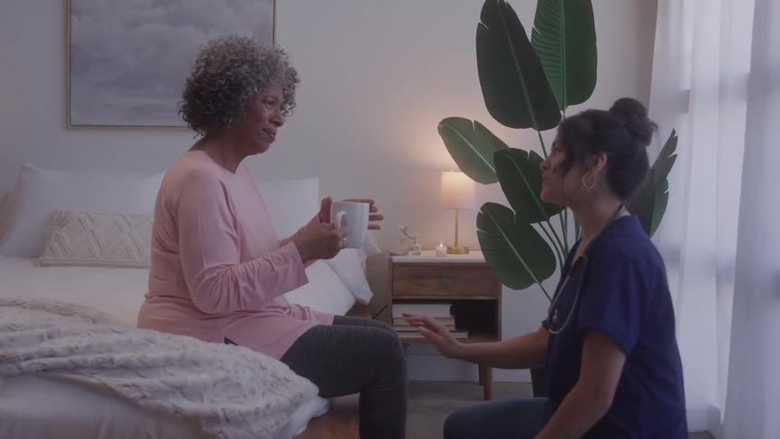 Woman sitting on the bed holding a cup while talking to a healthcare professional kneeling in front of her