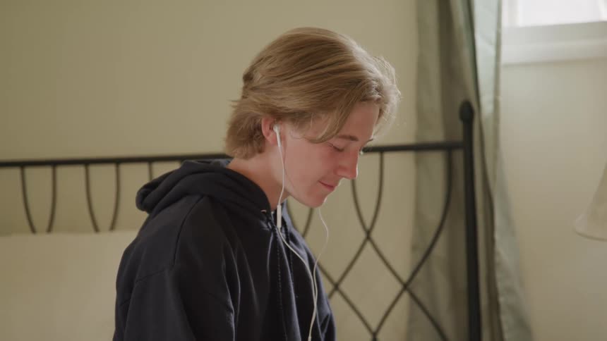 A close view of a teenager with light colored hair wearing a dark hoodie and white earphones and looking down in a bedroom