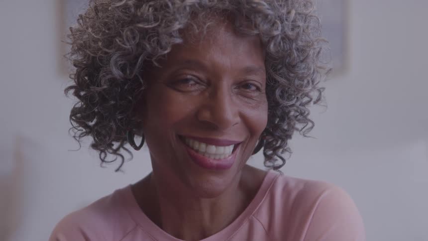A woman with gray and dark curly hair and a pink shirt is seen from a close view looking to the side and then directly at the camera with a wide smile on her face