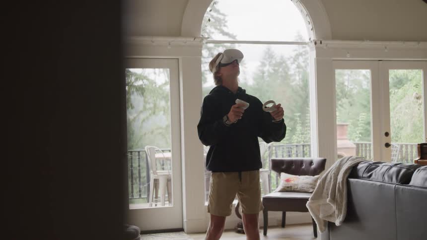 A woman carrying a backpack and a plastic bag walks into a sunfilled room with large glass doors that open to a patio she is wearing a backward baseball cap and appears to be enjoying the warm weather