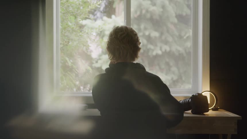 A person sits at a desk in front of a window with a digital camera nearby