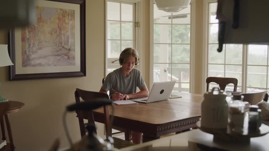A teenage male wearing headphones sits at his laptop and takes notes