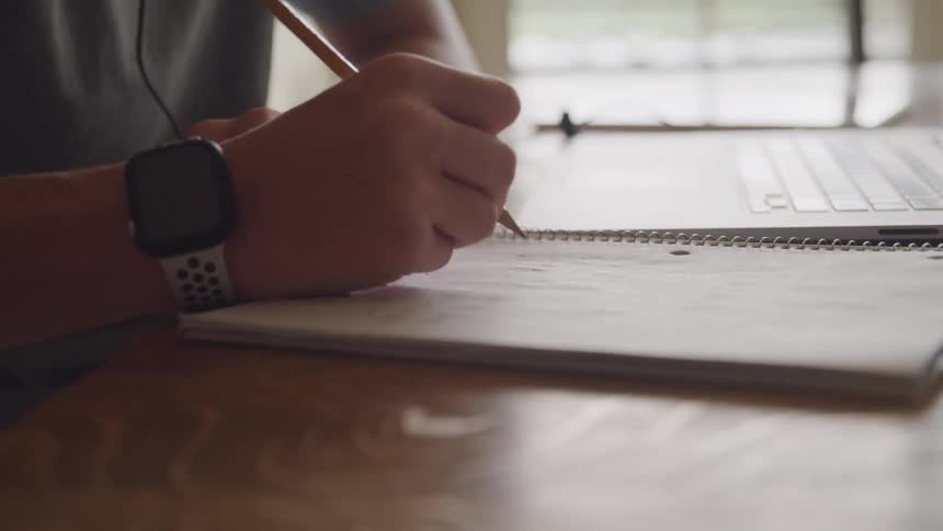 Female student writes in a notebook set on the table near a laptop during the daytime