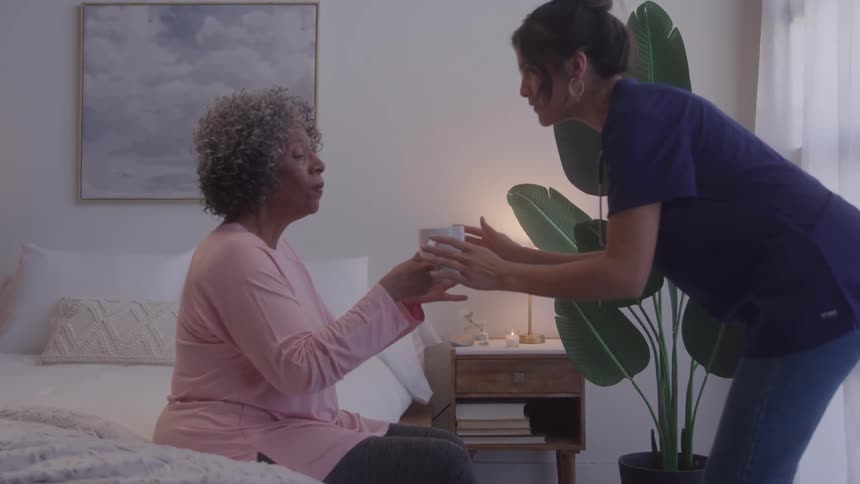 A female patient with gray hair is sitting on a bed and is handed a mug by a female medical staff member who is wearing dark blue scrubs and a stethoscope