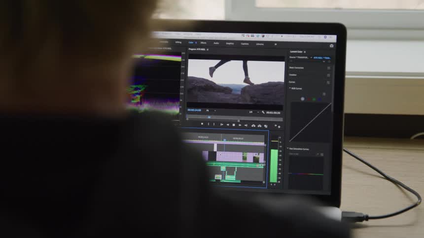 An editor works on a laptop timeline arranging clips and color curves