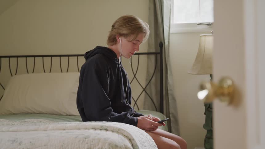 A person with earphones looks at their phone on a bed in a bedroom