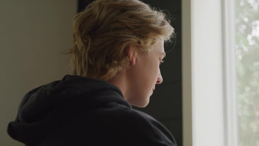 Young blondehaired person wears a black hoodie while looking out of a bright window