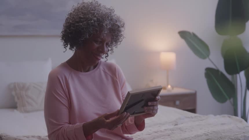 Woman holds a picture frame close to her chest in a bedroom softly lit by a lamp