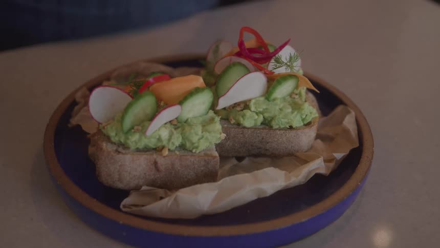 Fresh avocado toast is finished with colorful toppings as a hand carefully plates the dish