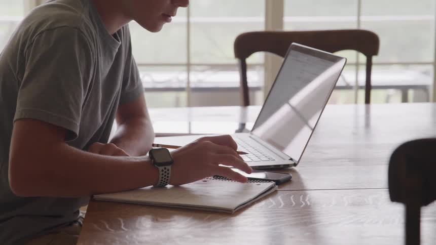 The video shows a person sitting at a wooden table wearing a gray tshirt and a smartwatch they are actively engaged in work or study using a laptop a smartphone and a notepad with a pencil