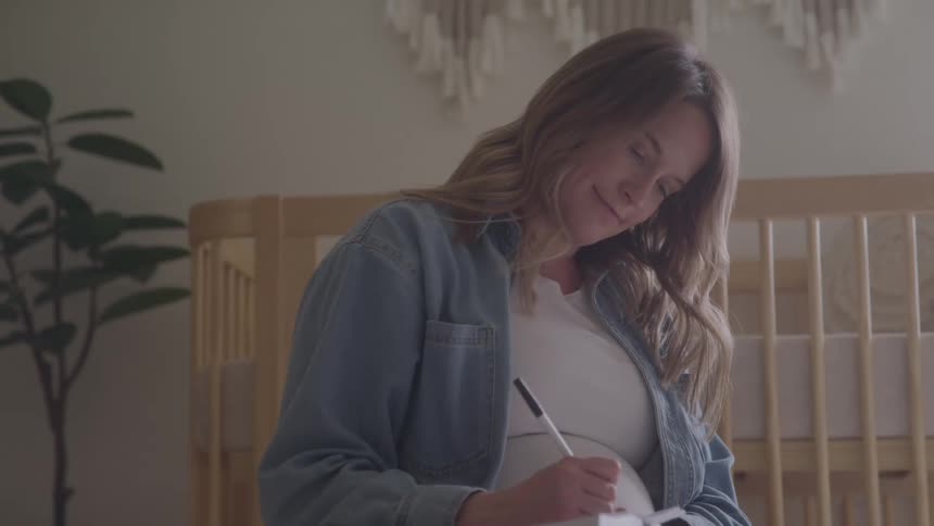 Pregnant woman writes in a notebook beside a crib