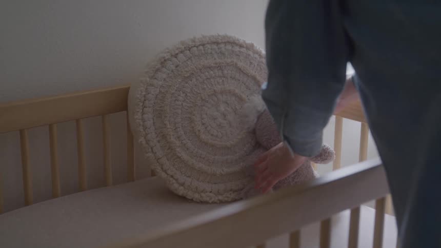 A woman arranges a cushion and teddy bear in a baby crib inside a room during daytime