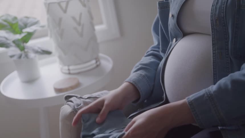 A close view of a pregnant woman sitting in a chair shows her folding a small blue baby garment near her large belly