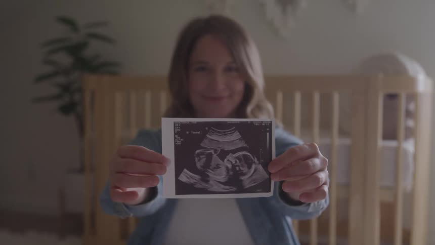 A pregnant woman displays her pregnancy ultrasound photo while sitting down in a room during daytime