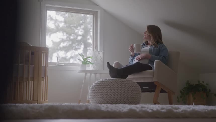 Pregnant woman sitting in an armchair with a mug in hand touching her belly in a room lit by natural light