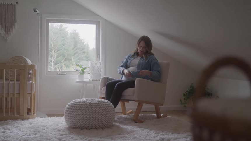 The video shows a pregnant woman sitting in a rocking chair in a babys nursery she is holding up a small gray baby garment and smiling