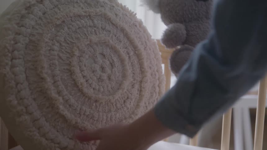 A close view shows a hand placing a beige textured pillow and a small grey stuffed koala with a white fuzzy nose into a light colored wooden crib with a white railing