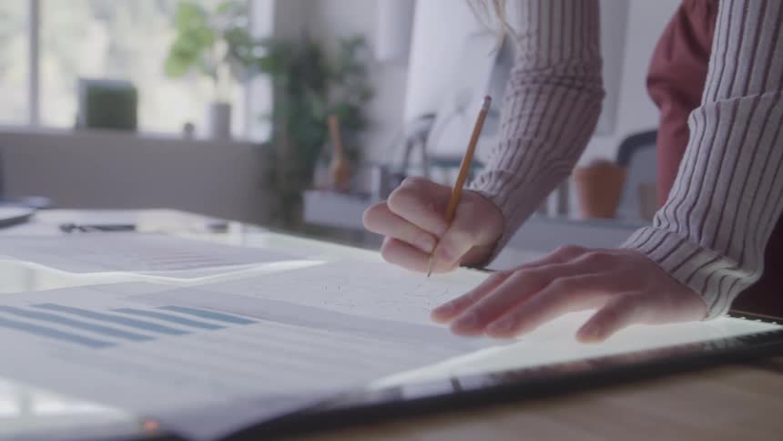 The video shows a closeup shot of a persons hands and torso as they lean over a table and work on a document they are using a light box to trace or draw on a paper with a pencil