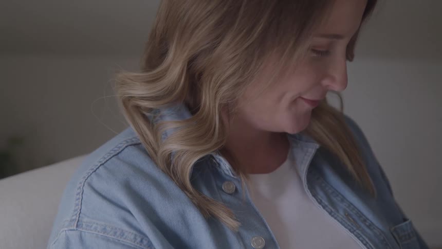 The video shows a pregnant woman with long blonde hair smiling and looking down she is wearing a white shirt and a blue denim jacket