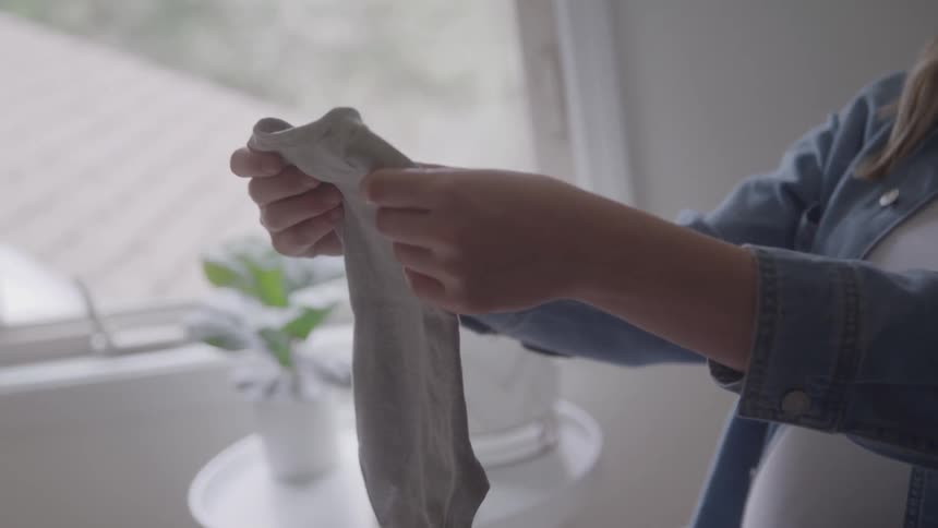 A pregnant woman folds baby clothes while sitting in a softly lit room