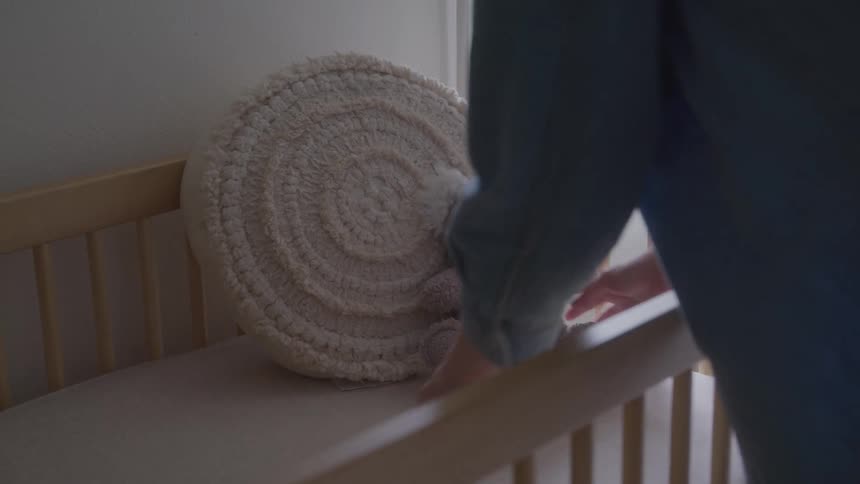 A person places a large beige pillow with a circular pattern and a small gray stuffed koala into a wooden crib