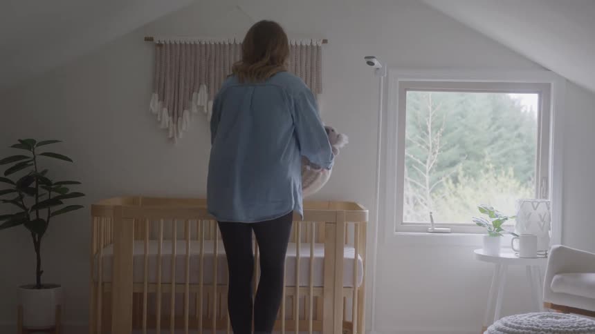A pregnant woman walks toward a wooden crib and bends down to place a stuffed animal and a small blanket inside