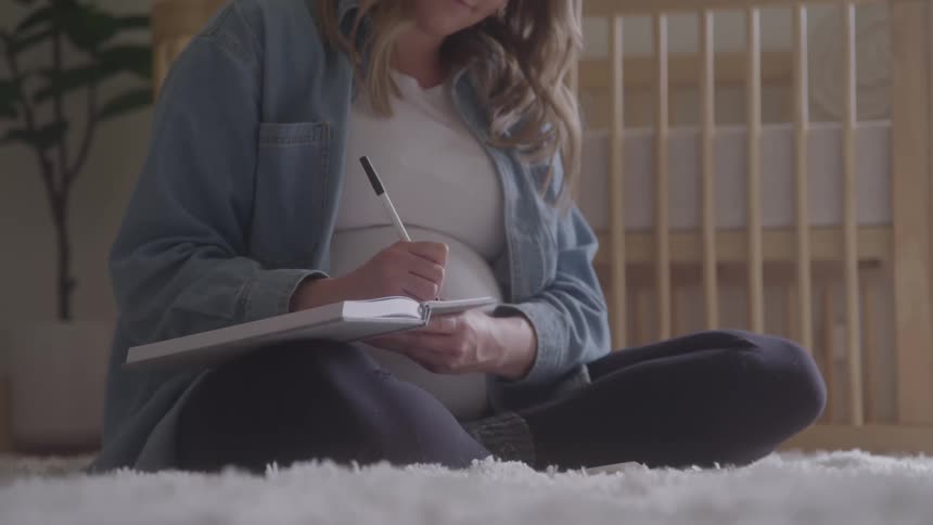 Pregnant woman sitting on rug next to crib writing in a journal