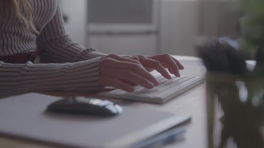 A close view of a persons hands and arms shows them typing on a white computer keyboard on a table with a wireless mouse and a stack of papers nearby
