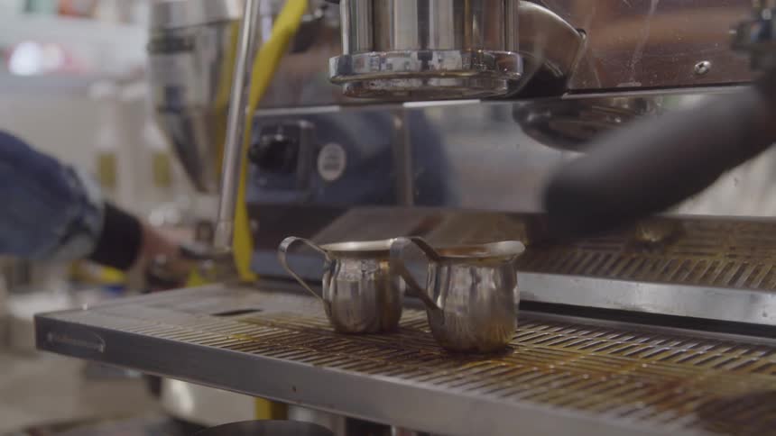 Two small metal pitchers rest on the drip tray of an espresso machine ready to catch freshly brewed coffee
